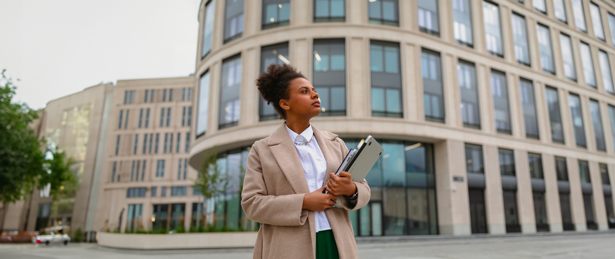 Frau mit Laptop und Kalender im Arm steht vor einem Bürogebäude